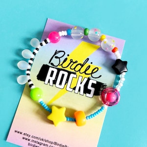 Puede incluir: Un brazalete de cuentas de colores con una variedad de encantos, incluyendo estrellas, una cuenta transparente con remolinos rosa y blanco, y una cuenta transparente con puntos blancos. El brazalete está en una tarjeta con el texto "Birdie ROCKS" y una dirección de sitio web.