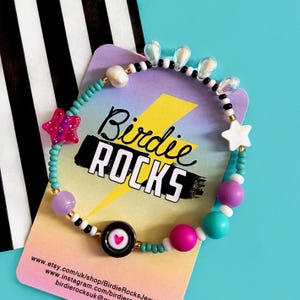 May include: A colorful beaded bracelet with a variety of beads, including stars, pearls, and round beads in pink, purple, and turquoise. The bracelet is displayed on a card with the text "Birdie ROCKS".