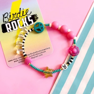 May include: A turquoise, pink and gold beaded bracelet with a peace sign charm, a star charm and a Taurus zodiac sign charm. The bracelet also features clear teardrop beads and black and white beads.