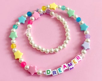 Può includere: Un braccialetto di perline color pastello con un braccialetto di perle sovrapposto. Il braccialetto di perline ha perline a forma di stella e perline con lettere che compongono la parola "DREAMER".
