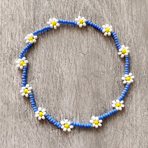 May include: A blue beaded bracelet with white daisy flower beads. The bracelet is made with small, round beads and features a repeating pattern of daisy flowers.