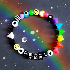 May include: A colourful beaded bracelet with black letter beads spelling out "REBEL" and various other colourful beads including smiley faces, stars, hearts, and spikes.