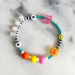 May include: A turquoise beaded bracelet with colorful beads, a pink star charm, a white heart bead, a white star bead, a yellow smiley face bead, and a clear bead with a yellow center.