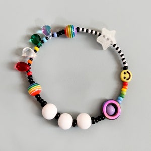 Op de afbeelding: Een kleurrijke kralen armband met witte, zwarte, regenboog en gele kralen. De armband heeft een ster bedel, een smiley bedel en een paarse en witte kraal met een kleine witte kraal erin.