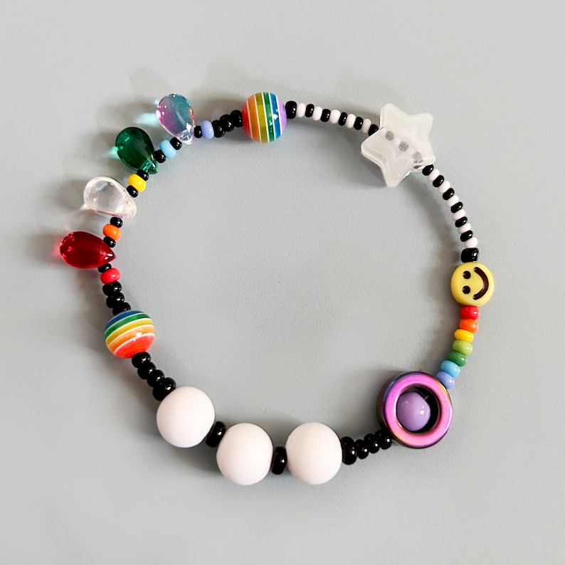 Op de afbeelding: Een kleurrijke kralen armband met witte, zwarte, regenboog en gele kralen. De armband heeft een ster bedel, een smiley bedel en een paarse en witte kraal met een kleine witte kraal erin.