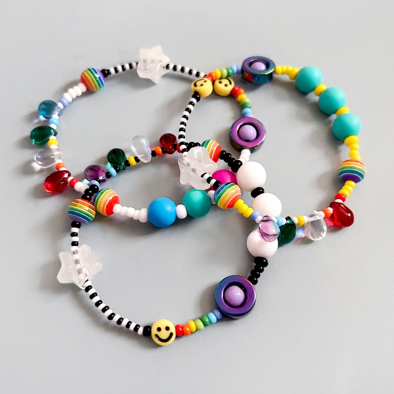 Op de afbeelding: Een kleurrijke kralenarmband met regenboog-, witte, zwarte en turquoise kralen. De armband heeft ster-, smiley- en cirkelcharms.