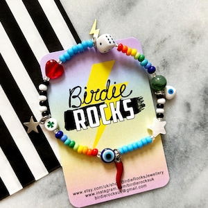 May include: A colorful beaded bracelet with charms, including a dice, a heart, a four-leaf clover, a star, an evil eye, and a chili pepper. The bracelet is on a card with the text "Birdie Rocks" and website information.