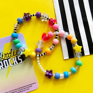 Könnte beinhalten: Zwei bunte Perlenarmbänder mit Regenbogenperlen, weißen Perlen und Stern- und Herzanhängern. Die Armbänder liegen auf einer gelben Oberfläche mit einer Visitenkarte, auf der "Birdie Rocks" steht.