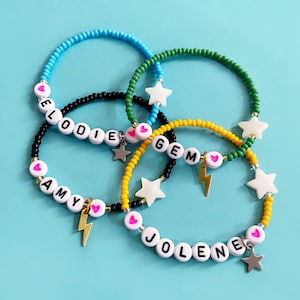 May include: Four colorful beaded bracelets with white letter beads spelling out the names "Elodie", "Amy", "Gem", and "Jolene". Each bracelet has a pink heart charm and a star charm.