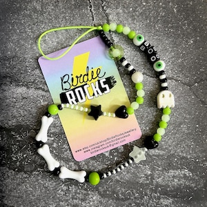 Puede incluir: Una pulsera de cuentas colorida con un cordón verde neón. La pulsera presenta cuentas negras, blancas y verdes, incluyendo cuentas con forma de hueso, estrella, corazón y fantasma. También hay una tarjeta de visita con el texto "Birdie Rocks".