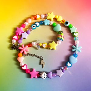 May include: A vibrant beaded necklace with star, heart, and flower-shaped beads. The necklace includes a silver chain and a lightning bolt charm. The beads are in pink, yellow, blue, and purple hues.