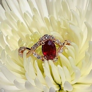 May include: A red ruby gemstone ring set in a rose gold band with a diamond halo. The ring is on a white flower.