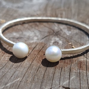 May include: A silver bangle bracelet with two white pearl accents. The bracelet is hammered and has a rustic look.