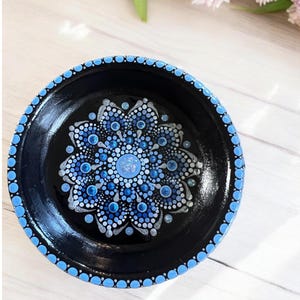 May include: A black circular dish with a blue and white mandala design painted on the surface. The mandala is made up of small dots and circles.