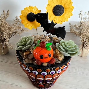 Puede incluir: Una pequeña maceta de terracota con un patrón de lunares negros y naranjas está llena de pequeñas piedras, dos pequeñas suculentas verdes y una pequeña figurita de calabaza naranja con una cara sonriente. Dos girasoles amarillos y un murciélago negro se colocan en la parte superior de la maceta.