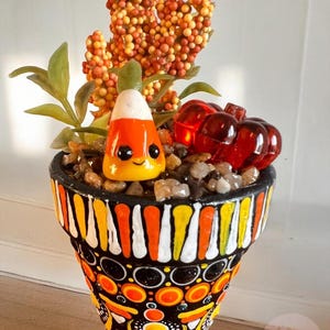 Puede incluir: Maceta decorativa pintada a mano con temática de Halloween. La maceta presenta una base negra con patrones naranjas, amarillos y blancos. Contiene una figura de caramelo de maíz, piedras decorativas y follaje artificial. También se incluyen dos decoraciones de calabaza roja.