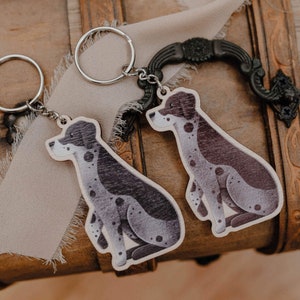 May include: Two wooden dog keyrings with silver rings. The dogs have black and grey spotted markings on a light wood background. The keyrings are on a wooden chest with a beige fabric.