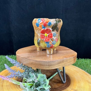 May include: A small, ceramic flower pot with a floral design. The pot is tan with a large red flower and smaller blue, orange, and yellow flowers. It sits on a small wooden stand with metal legs. The background is black.