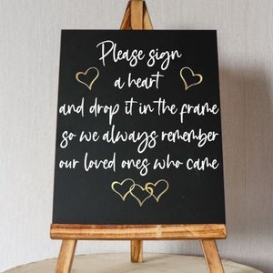 以下が含まれることがあります： 木製のイーゼルが付いた黒いチョークボードのサインで、「心をサインしてフレームに入れてください。そうすれば、来た人々をいつも忘れません」というテキストが書かれており、金色のハートのアクセントが付けられています。