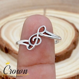 May include: A silver ring with a treble clef design. The ring is worn on a finger.