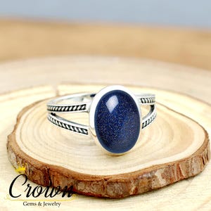 May include: A silver ring featuring a large, oval, dark blue gemstone with a speckled appearance. The ring has a split band design with decorative detailing. The ring is displayed on a wooden surface.