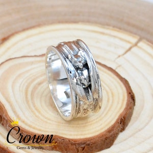 May include: A silver ring with a textured design featuring a small animal figure. The ring is wide and has a polished finish.