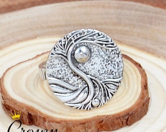 May include: A silver ring with a textured, circular design featuring a tree and a wave. The ring is on a wooden surface.