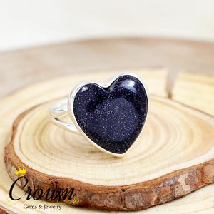 Puede incluir: Un anillo de plata con una piedra preciosa azul en forma de corazón. La piedra preciosa tiene una textura brillante y moteada.