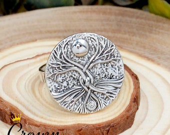 May include: A silver ring with a tree of life design. The ring features a detailed, textured design of a tree with branches reaching up to a circular moon. The ring is on a wooden surface.