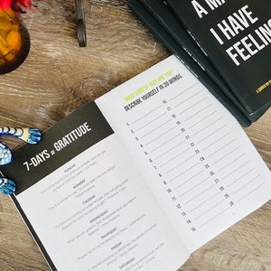 Pu&ograve; includere: Un diario aperto con una copertina nera e pagine bianche. Il diario ha una sezione intitolata "7-Days of Gratitude" e un'altra sezione intitolata "What Kind of Man Are You? Describe Yourself in 30 Words." Il diario &egrave; aperto a una pagina con un elenco dei giorni della settimana e suggerimenti per ogni giorno. Il diario &egrave; su una superficie di legno.