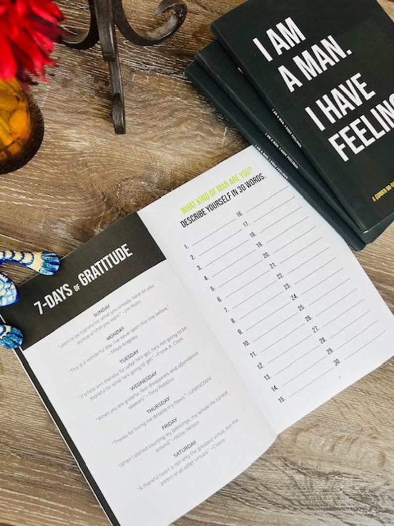 Pu&ograve; includere: Un diario aperto con una copertina nera e pagine bianche. Il diario ha una sezione intitolata "7-Days of Gratitude" e un'altra sezione intitolata "What Kind of Man Are You? Describe Yourself in 30 Words." Il diario &egrave; aperto a una pagina con un elenco dei giorni della settimana e suggerimenti per ogni giorno. Il diario &egrave; su una superficie di legno.