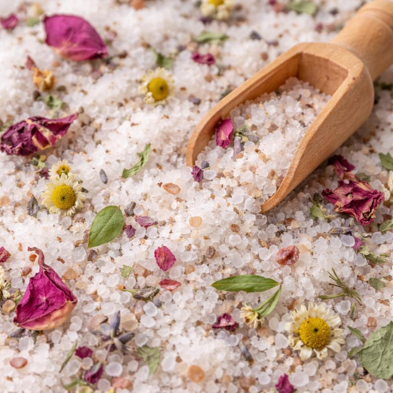 May include: Close-up of bath salts with dried rose petals, chamomile flowers, and a wooden scoop. The salts are a mix of white and tan granules, with green herbs and lavender buds scattered throughout. The wooden scoop is partially filled with the bath salts.
