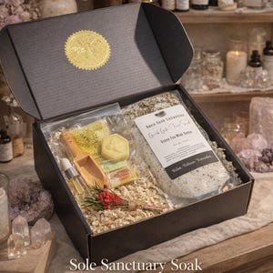 May include: A black gift box with a gold seal, filled with bath products. Includes a bag of Green Tea Mint Detox, a wooden scoop, a small bottle, and a bar of soap. The box is labeled "Sole Sanctuary Soak".