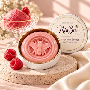 May include: A pink solid lotion bar embossed with a bee design, housed in a white metal tin with a lid labeled Honoring Miss Bee Raspberry Sorbet Solid Lotion Bar, accompanied by fresh raspberries and a scoop of raspberry sorbet.