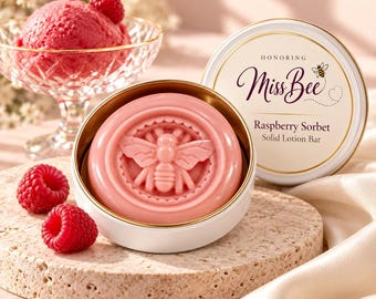 Może przedstawiać: Różowy stały balsam w kostce z wytłoczonym wzorem pszczoły, umieszczony w białej metalowej puszce z wieczkiem z napisem Honoring Miss Bee Raspberry Sorbet Solid Lotion Bar, w towarzystwie świeżych malin i sorbetu malinowego.