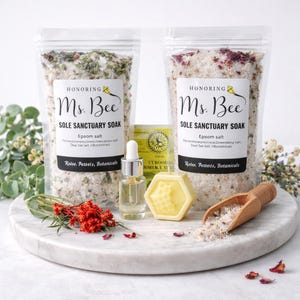 May include: Two clear bags of bath salts labeled "Ms. Bee Sole Sanctuary Soak" with Epsom salt, and botanicals. A small bottle of oil, a yellow honeycomb-shaped soap, and a wooden scoop with bath salts are on a marble tray.
