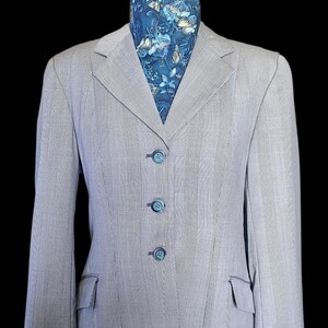 Puede incluir: Una blazer gris claro con un sutil estampado de rayas, tres botones a juego y dos bolsillos delanteros. Una bufanda estampada con diseños florales azules y blancos es visible en el escote. La blazer se exhibe en un maniquí negro.