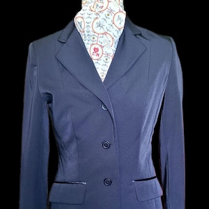 May include: A navy blue blazer with a two-button closure and two front pockets. The blazer has a contrasting black trim on the pockets and lapels. A patterned scarf with red and white accents is visible at the neckline.