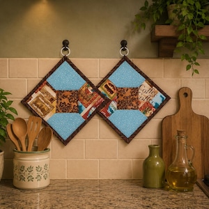 May include: Two quilted pot holders with a blue and brown patchwork design, hanging on black hooks. The pot holders have a diamond shape with a brown border. The background includes a wooden cutting board, a green vase, and a glass bottle.