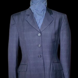 Puede incluir: Chaqueta de equitación azul marino con un sutil estampado de cuadros. La chaqueta tiene un cuello de terciopelo negro, tres botones y dos bolsillos delanteros. Se lleva un pañuelo azul y blanco con lunares debajo.
