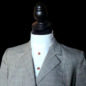 Op de afbeelding: Een grijze geruite colbertjas met een wit overhemd en bruine knopen. De jas wordt getoond op een zwarte mannequinhoofd tegen een zwarte achtergrond. De colbertjas heeft een klassiek, op maat gemaakt ontwerp.