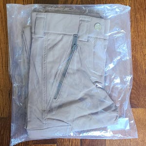 以下が含まれることがあります： 左足にジッパー付きポケットが付いた薄い茶色のパンツ。パンツは折り畳んで透明なビニール袋に入っています。
