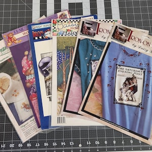 May include: A collection of six Daisy Kingdom iron-on transfer packets featuring various designs, including a Saturday Evening Post illustration, floral patterns, and teddy bears. Each package displays the product name and visual examples.