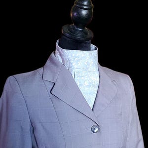 Puede incluir: Un blazer gris con un sutil patrón de cuadros, abotonado en la parte delantera. Se combina con una camisa de cuello azul claro con un diseño floral blanco. El conjunto se exhibe en un maniquí negro.