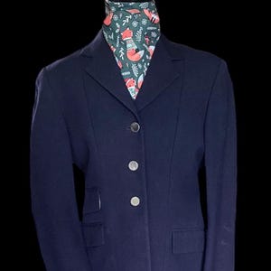 Pode incluir: Uma jaqueta de equitação azul marinho com botões prateados. A jaqueta tem gola e fecho de um único botão. A jaqueta está em um manequim que está usando um lenço estampado em verde e branco com raposas.