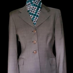 May include: A tailored, taupe-colored blazer with three buttons and two front pockets. A patterned scarf in shades of green, white, and brown is visible at the neckline. The jacket is displayed on a black mannequin.