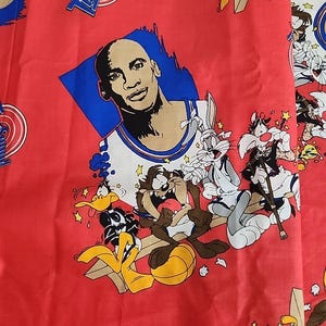 May include: Red fabric featuring a graphic of Michael Jordan and Looney Tunes characters. The design includes Bugs Bunny, Daffy Duck, and Taz, with the "Tune Squad" logo. The fabric has a repeating pattern of cartoon characters.