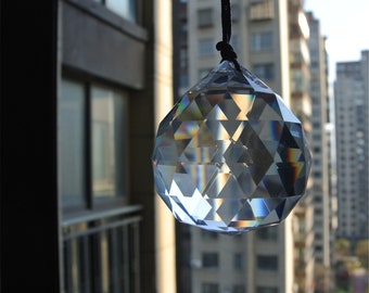 Puede incluir: Una bola de cristal transparente colgando de una cuerda negra frente a una ventana con vista a un horizonte urbano. La bola de cristal es facetada y refleja la luz.