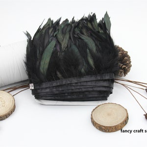 May include: Black feather trim with a green iridescent sheen. The trim is rolled up and tied with a black ribbon. The trim is on a white surface with wooden accents.