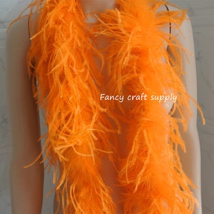 Op de afbeelding: Een feloranje verenboa. De boa is lang en pluche, en is over de schouders van een mannequin gehangen. De tekst "Fancy craft supply" is zichtbaar in de afbeelding.